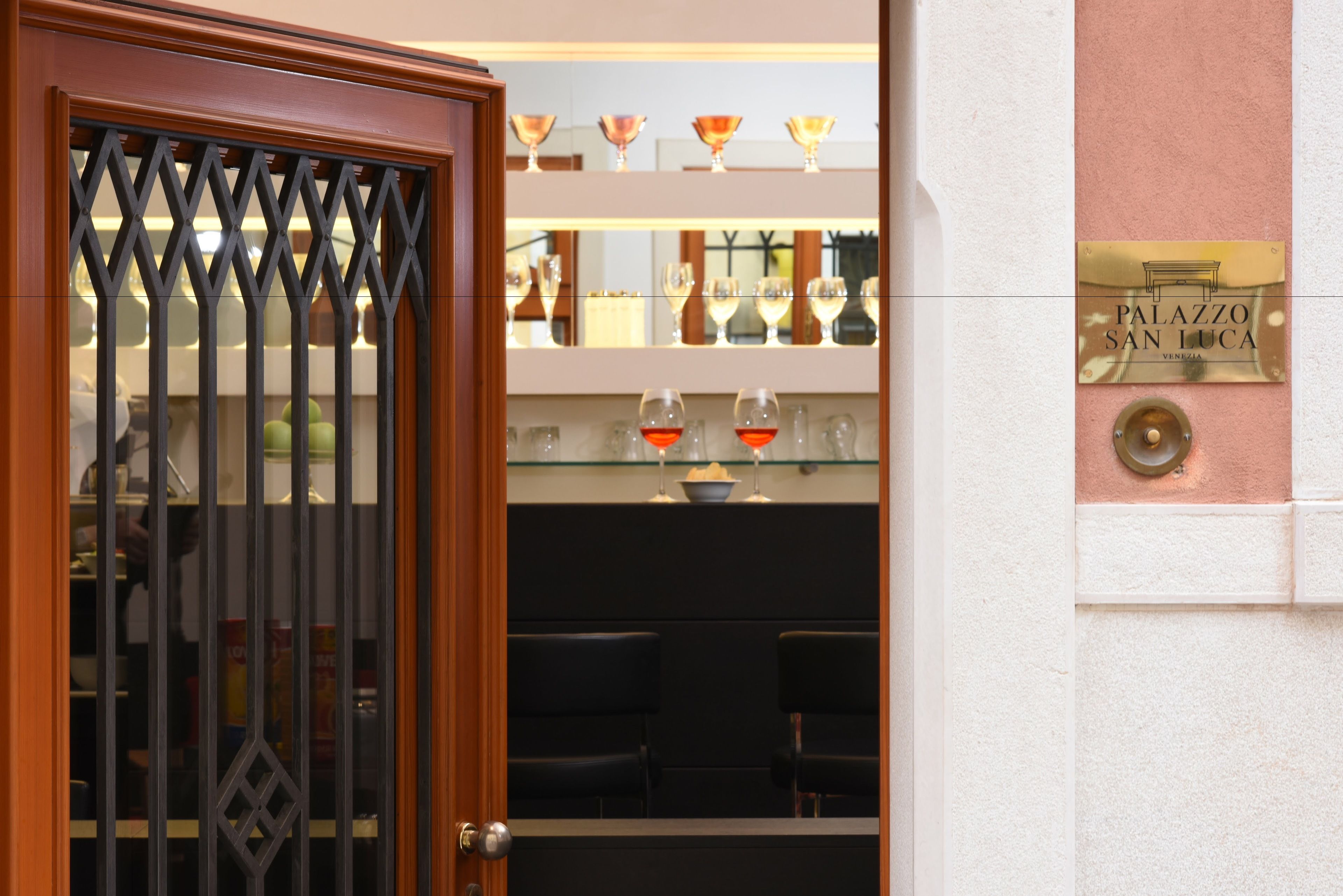 Entrada de un hotel con una reja metálica decorativa, una vitrina con copas de vino y una placa que dice "PALAZZO SAN LUCA VENEZIA".