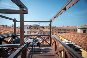Un balcon en bois, agrémenté d’une table et de chaises, d’où l’on peut admirer un paysage urbain avec des toits recouverts de tuiles rouges et des bâtiments.