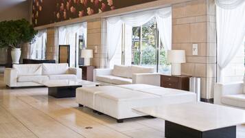 A spacious lobby with a modern design, featuring a large seating area with sofas and a coffee table, large windows with curtains, and a decorative wall with a leaf pattern.