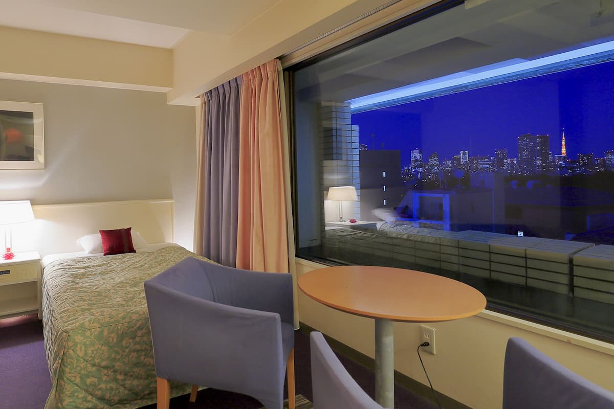 A hotel room with two beds, a nightstand with a lamp, a chair, a small table, and a large window offering a city view.