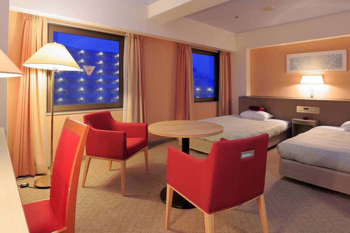 A hotel room with two beds, a desk, a chair, a small table, and a window with curtains.