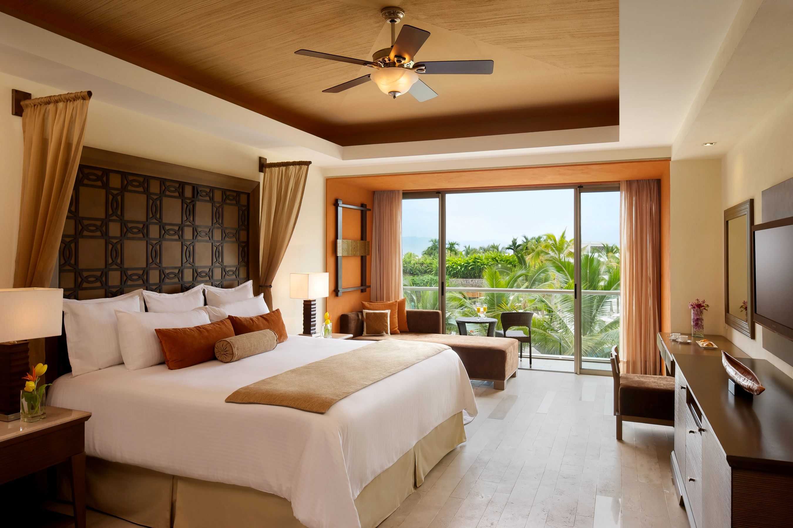 A hotel room with a large bed, a ceiling fan, a balcony with a view of palm trees, and a TV on the wall.