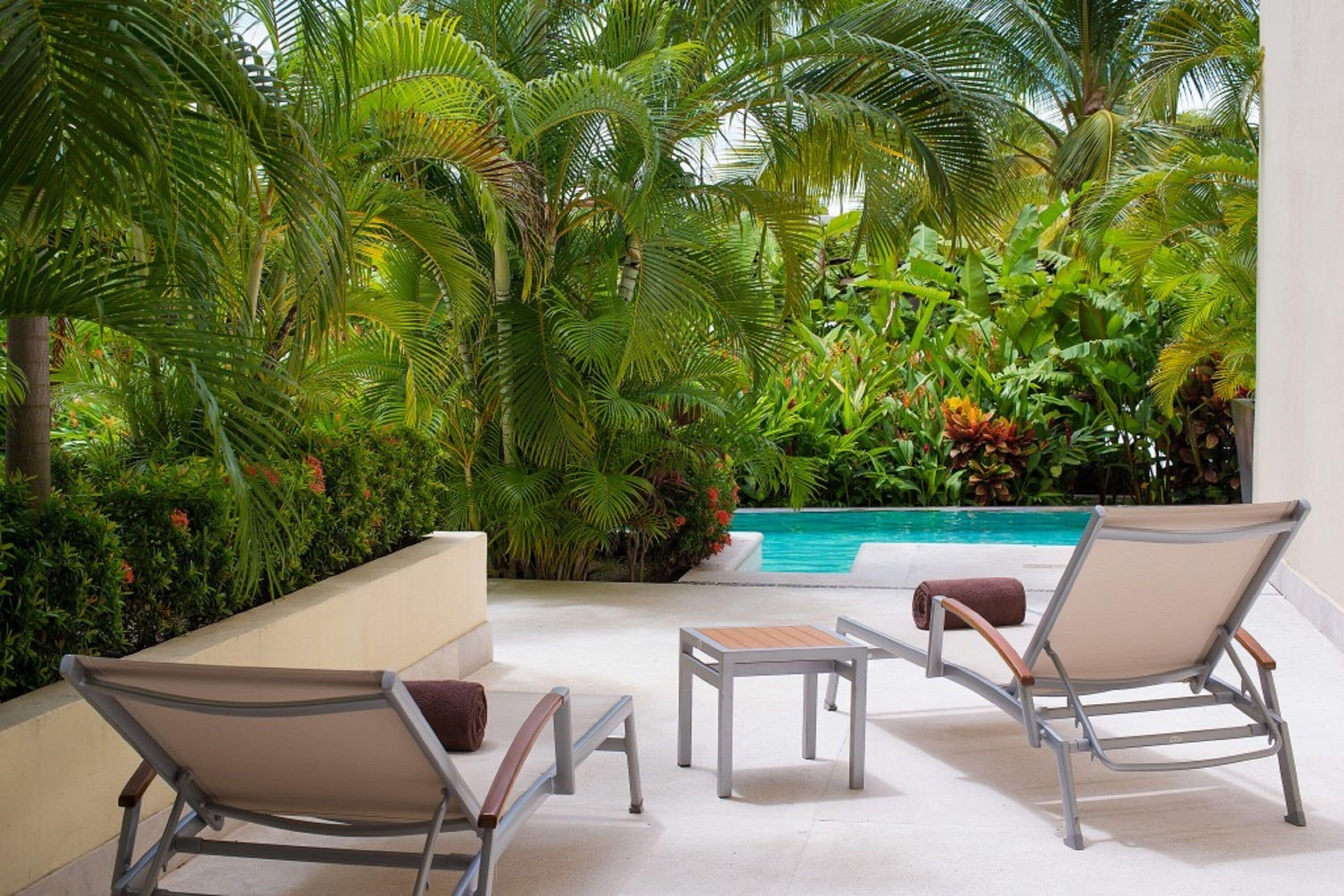 A poolside area with two lounge chairs and a small table, surrounded by lush tropical plants and a clear pool.