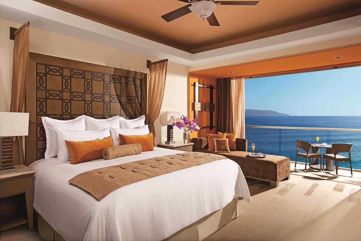 A hotel room with a large bed, a balcony with a table and chairs, and a view of the sea.