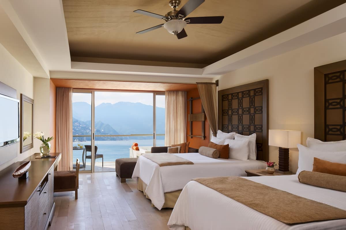 A hotel room with two beds, a desk, a chair, and a large window offering a view of the sea and mountains.