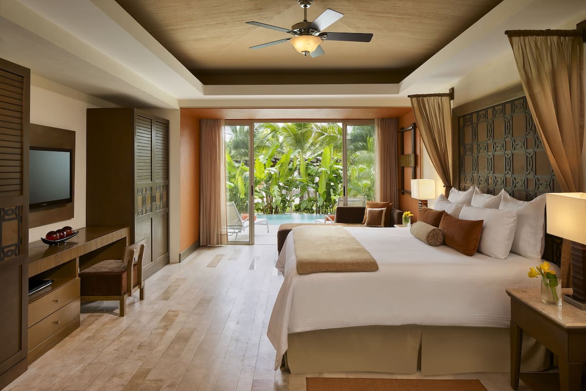 A hotel room with a bed, a TV, a desk, and a view of a pool and palm trees.