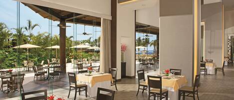 Un vaste espace de restauration intérieur, avec des tables dressées pour les clients, de grandes fenêtres offrant une vue sur les palmiers et la plage, et un design architectural moderne aux lignes épurées et aux couleurs neutres.