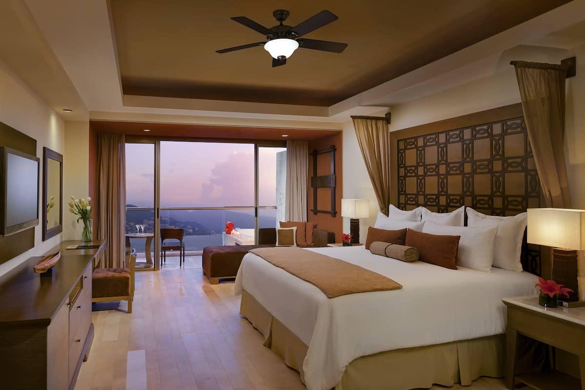 A spacious hotel room with a large bed, a ceiling fan, a balcony with a view, and a TV on the left side.