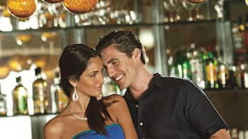Um casal em pé num bar, com luzes esféricas penduradas e garrafas ao fundo.