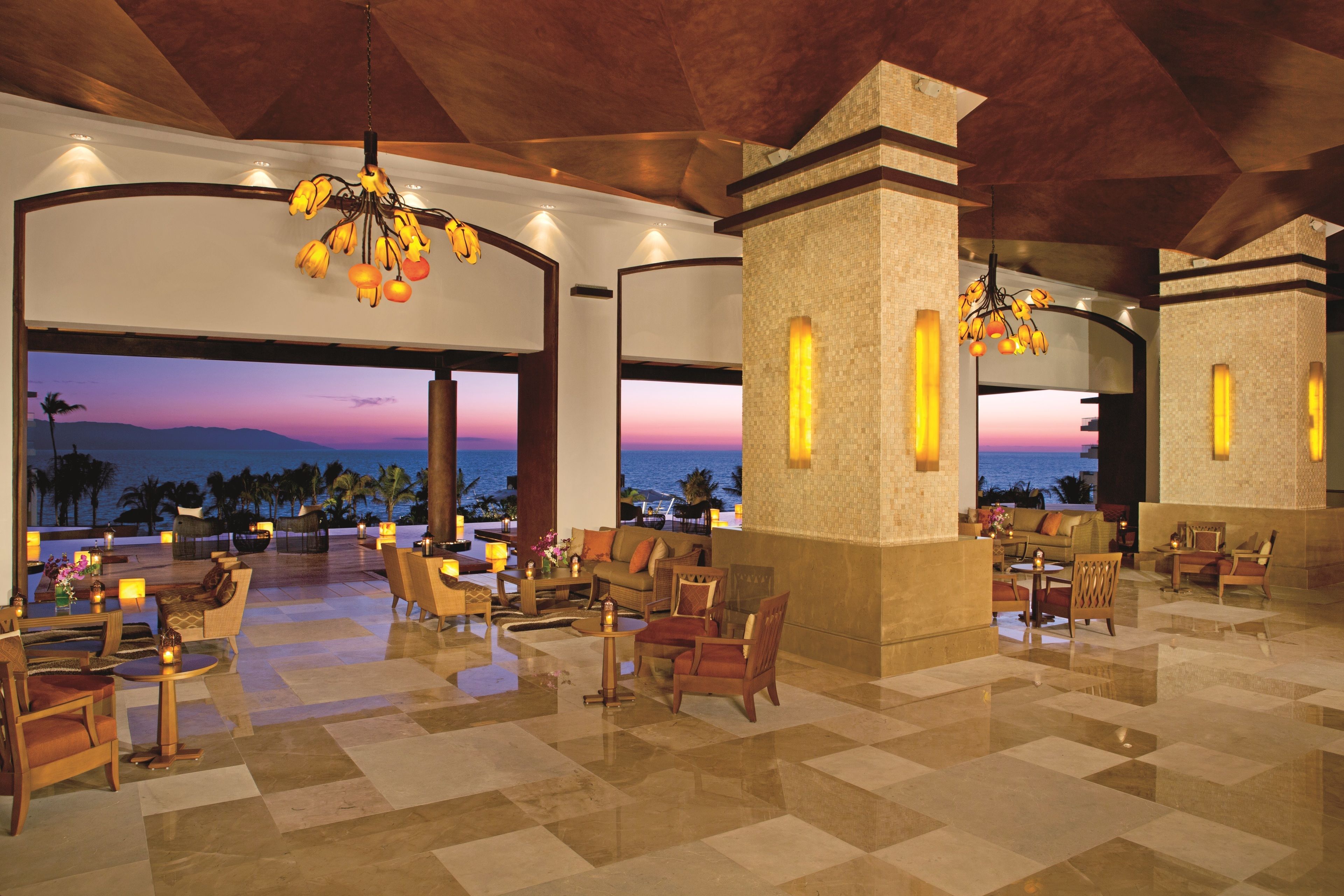 A spacious lobby with a view of the ocean at sunset, featuring comfortable seating and a modern chandelier.