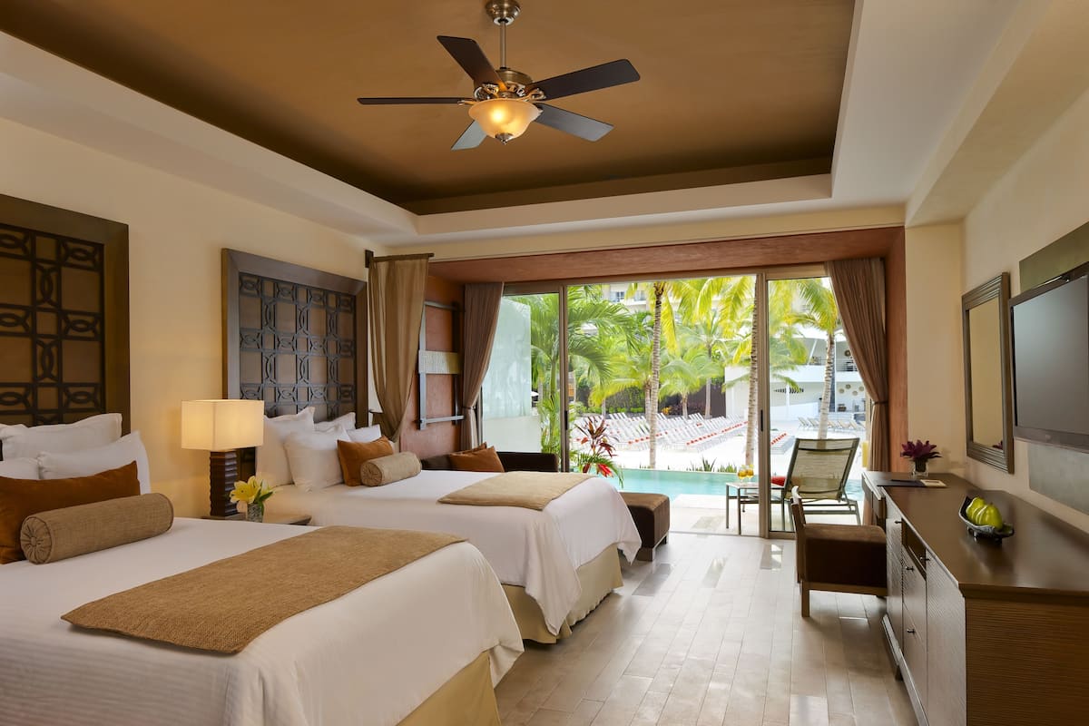A hotel room with two beds, a ceiling fan, a desk with a TV, and a view of a pool area.