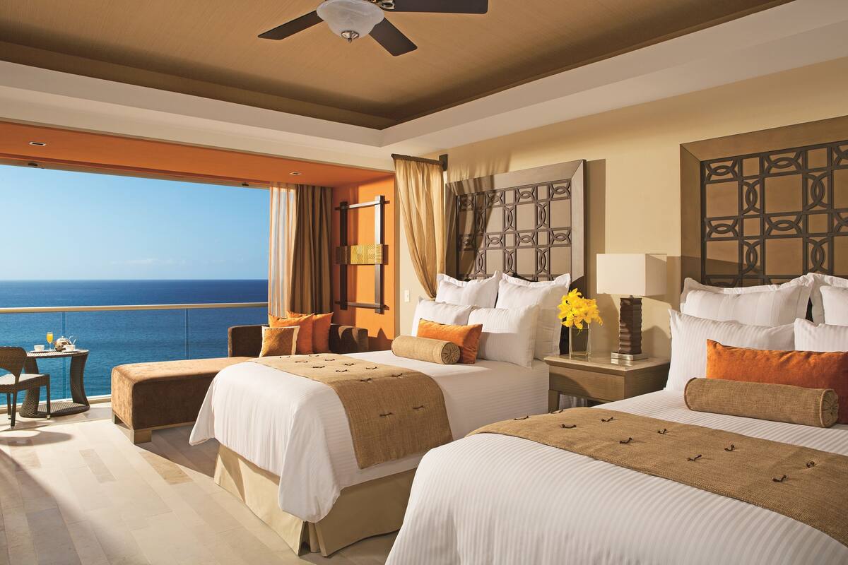 A hotel room with two beds, a balcony with a view of the sea, a ceiling fan, and a large decorative headboard.
