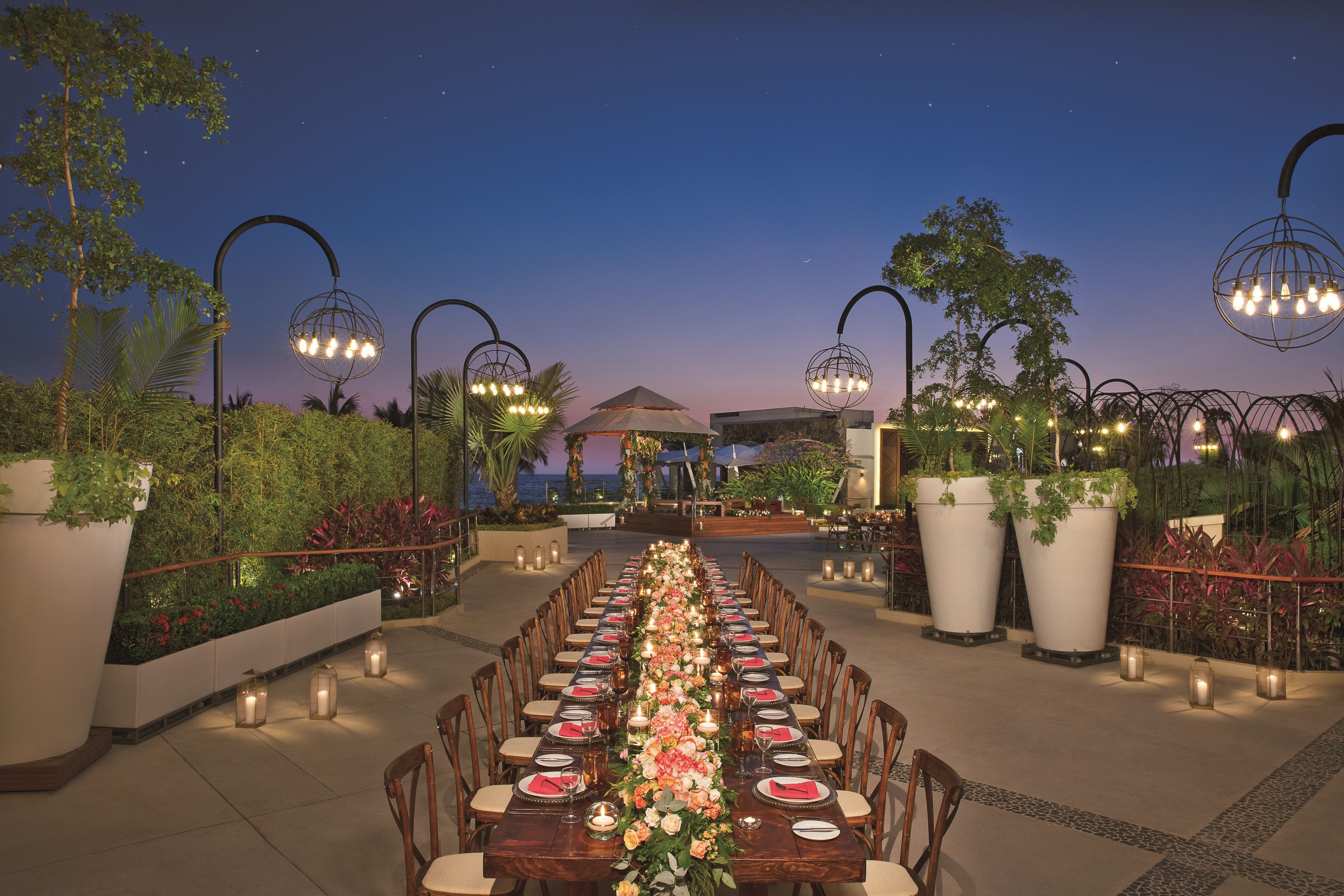 An outdoor dining area with a long table set for a meal, surrounded by potted plants and decorative lighting.