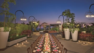 An outdoor dining area with a long table set for a meal, surrounded by potted plants and decorative lighting.