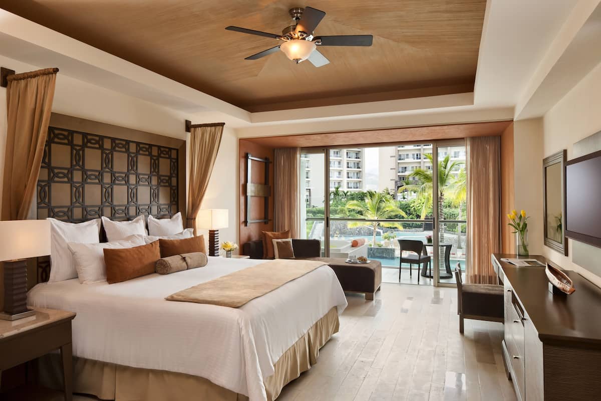 A hotel room with a large bed, a desk, a TV, and a balcony with a view of the city.