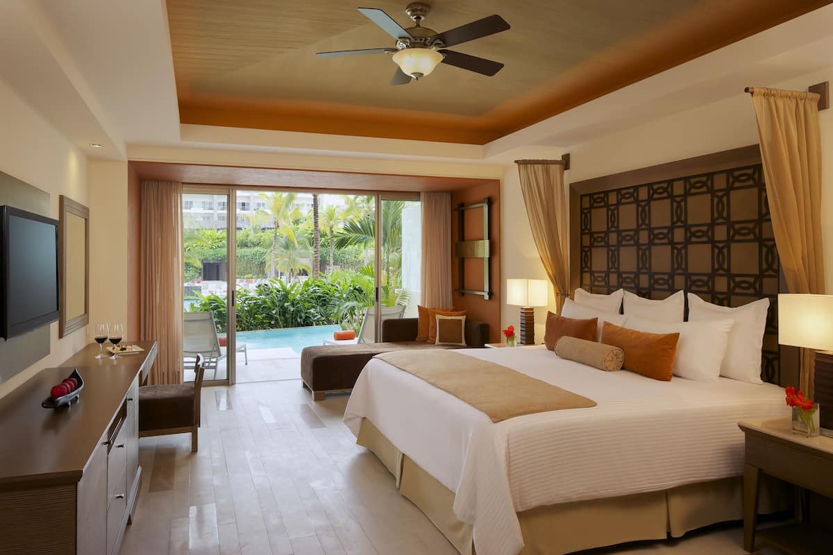 A hotel room with a large bed, a desk with a flat-screen TV, a balcony with a pool view, and a ceiling fan.