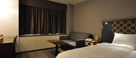 A hotel room with a bed, a desk, a chair, a small table, a telephone, and a window with curtains.