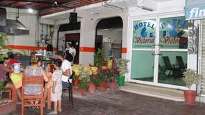 An outdoor seating area with tables and chairs, potted plants, and a visible entrance to a hotel named "Hotel Victoria". - Hotel Victoria Zihuatanejo (Zihuatanejo)