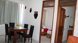 A dining area with a wooden table and chairs, a small flower vase, and a bed with a red blanket in the background. - Hotel Victoria Zihuatanejo (Zihuatanejo)