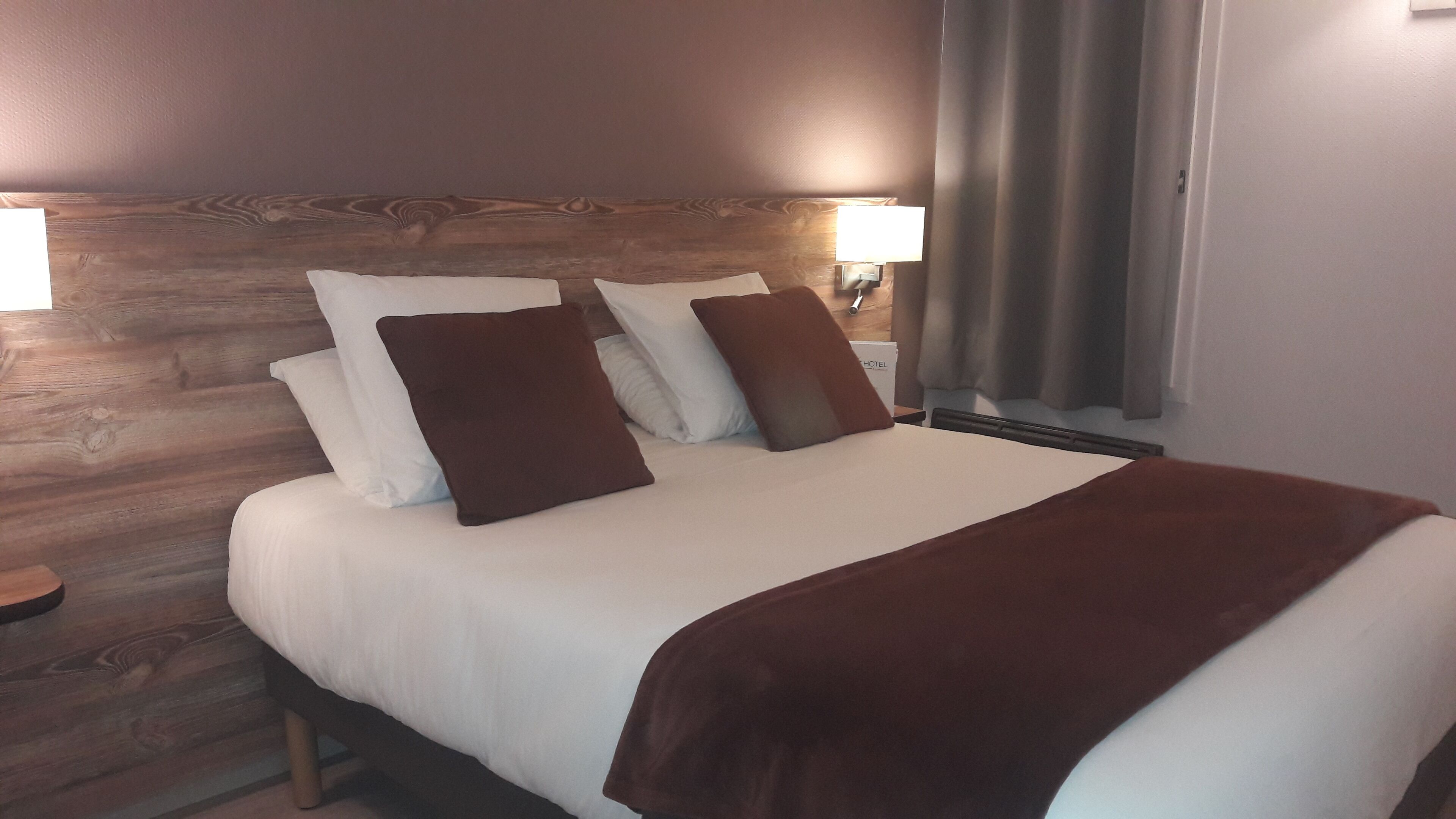 A hotel room with a wooden headboard, a large bed with white bedding and brown pillows, two bedside lamps, and a window with curtains.