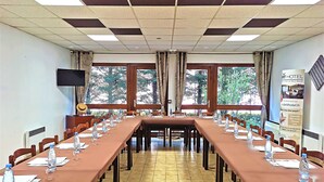 A conference room with long tables, chairs, and water bottles, large windows with a view of trees, and a flat-screen TV on the wall. - Brit Hotel Confort Pau Lons (Lons)