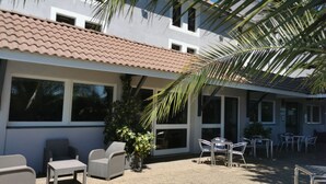 A building with a tiled roof, large windows, outdoor seating area with chairs and tables, and a palm tree. - Brit Hotel Confort Pau Lons (Lons)