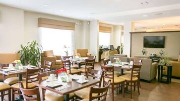 A hotel dining area with multiple tables and chairs, a sofa, and a television on the wall.