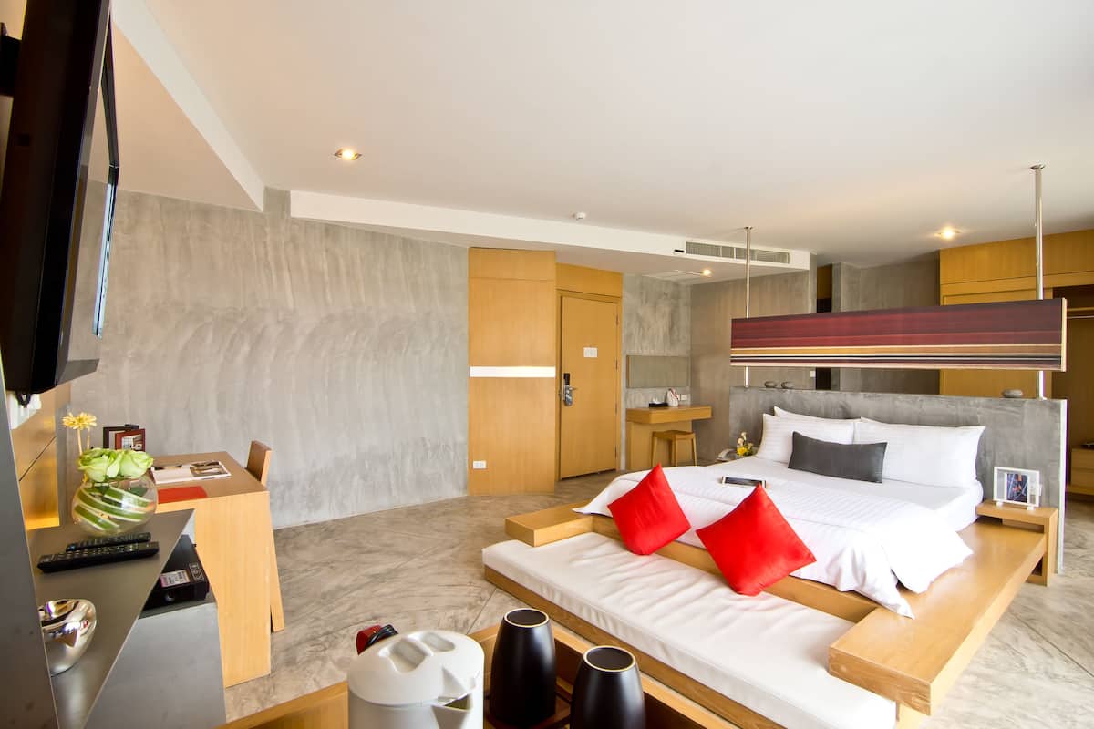 A modern hotel room with a large bed, wooden bedside tables, a desk with a chair, a TV, and a bathroom with a mirror and vanity.