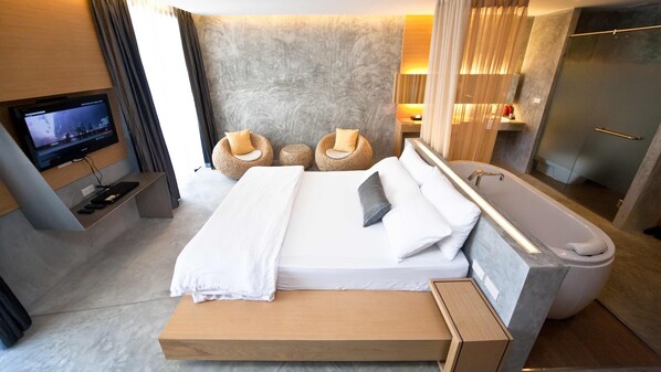 A modern hotel room with a large bed, a TV, a bathtub, and two round chairs.