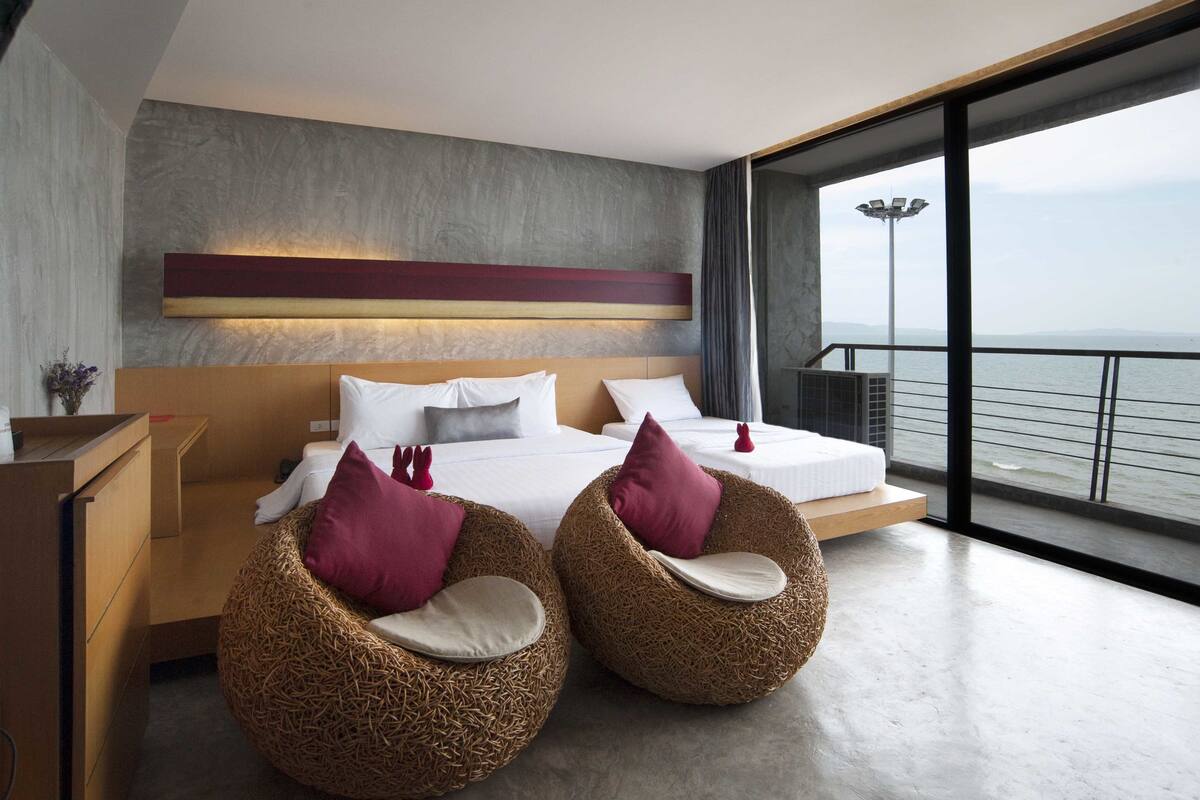 A modern hotel room with a large bed, two wicker chairs, a view of the sea, and a balcony.