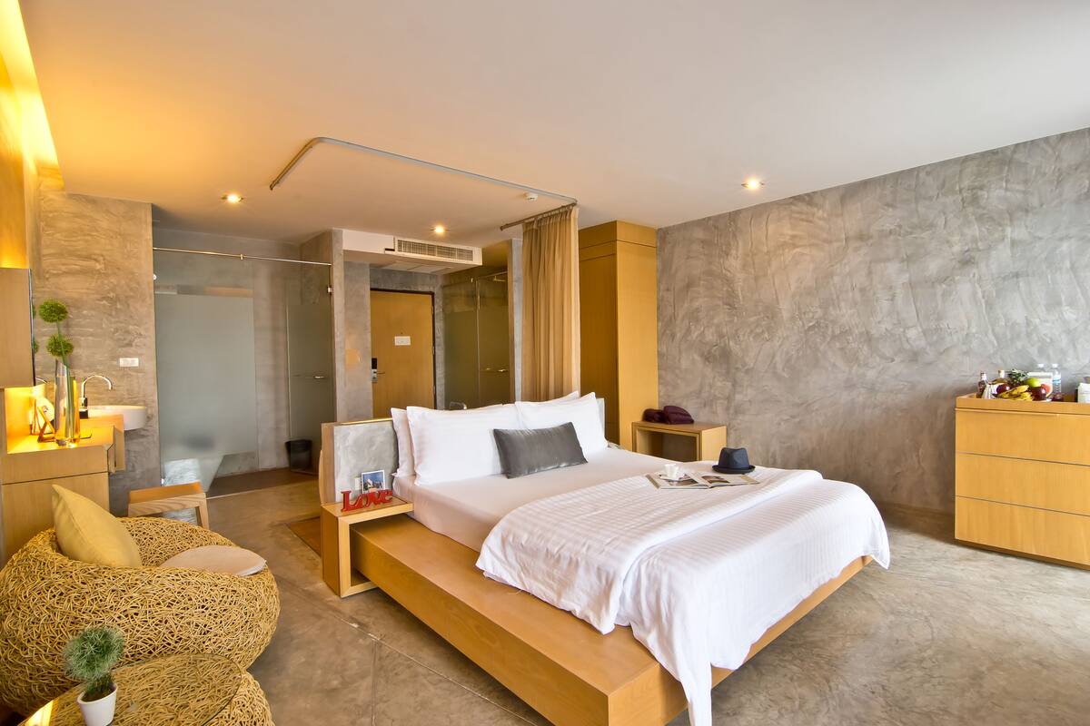 A modern bedroom with a large bed, bedside tables, a wooden bench, and a bathroom visible in the background.