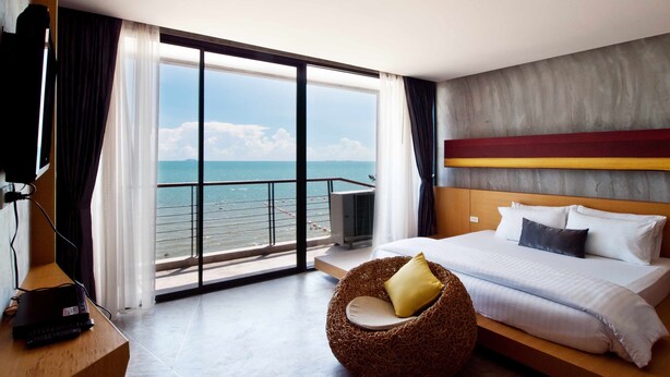 Habitació d'hotel amb una finestra gran que ofereix vista al mar, un llit amb roba de llit blanca, una cadira de veta i una televisió sobre una taula de fusta.