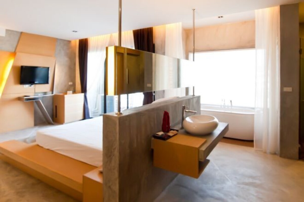 A modern hotel room with a large bed, a built-in vanity with a sink, a flat-screen TV mounted on the wall, and a large window providing natural light.