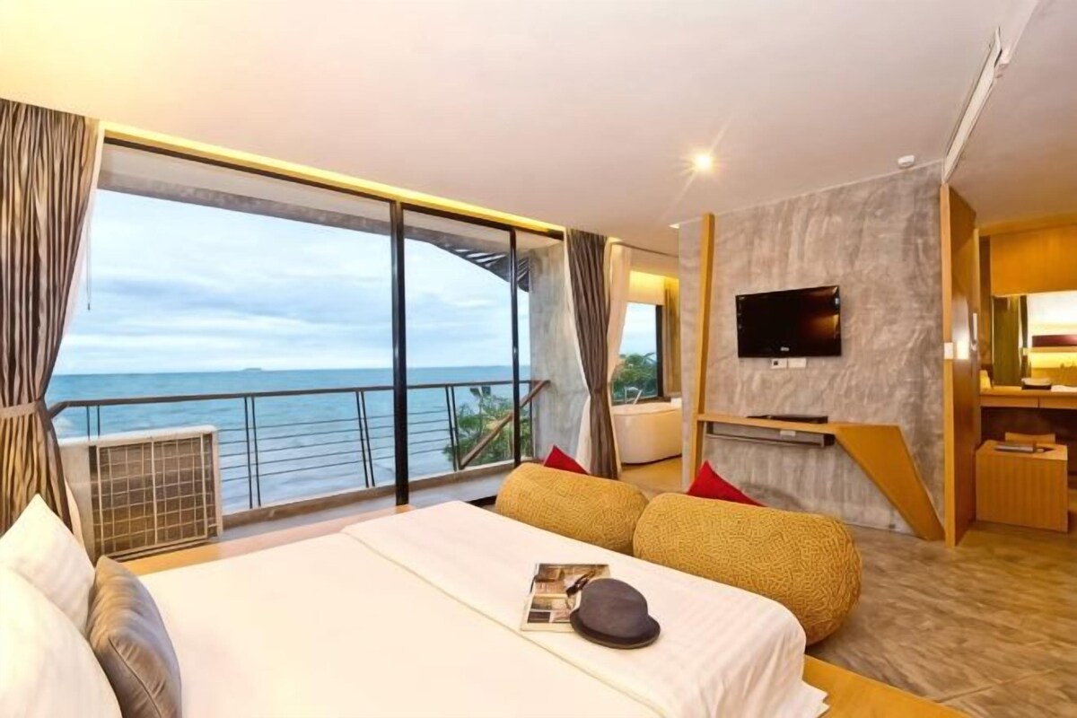 A hotel room with a large bed, a TV, a balcony with a view of the sea, and a small desk area.