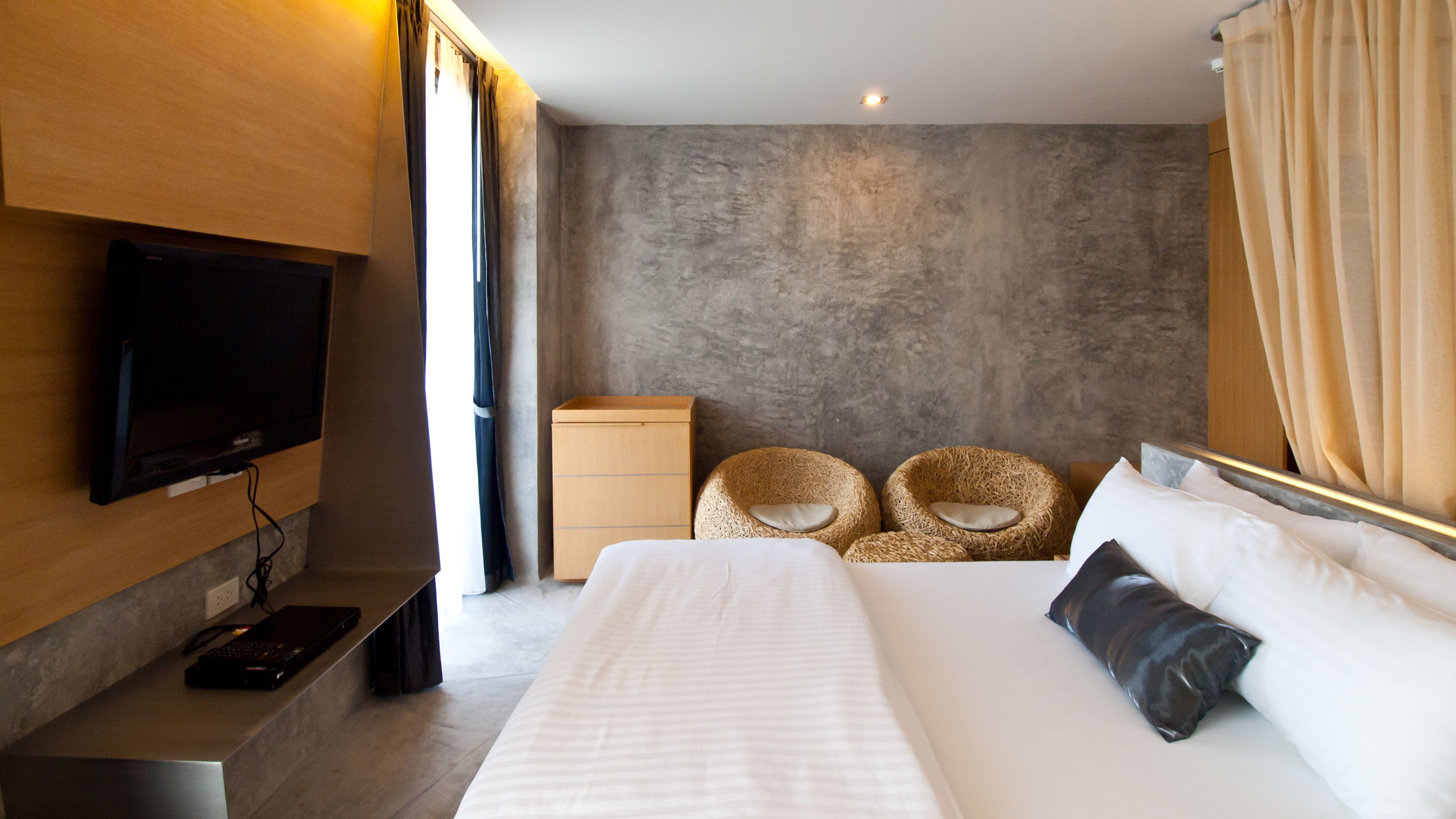 A modern hotel room with a large bed, a flat-screen TV mounted on the wall, a wooden nightstand, and two woven chairs.
