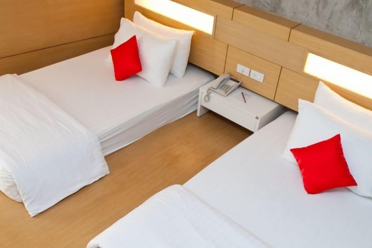 A hotel room with two single beds, each with white bedding and a red pillow. There is a bedside table with a phone and a lamp.