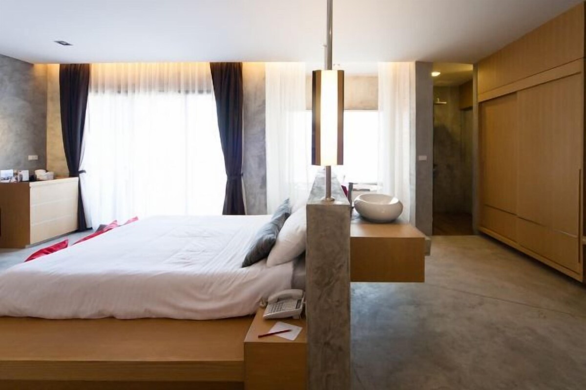 A modern hotel room with a large bed, bedside table, lamp, and a view of a cityscape through the window.