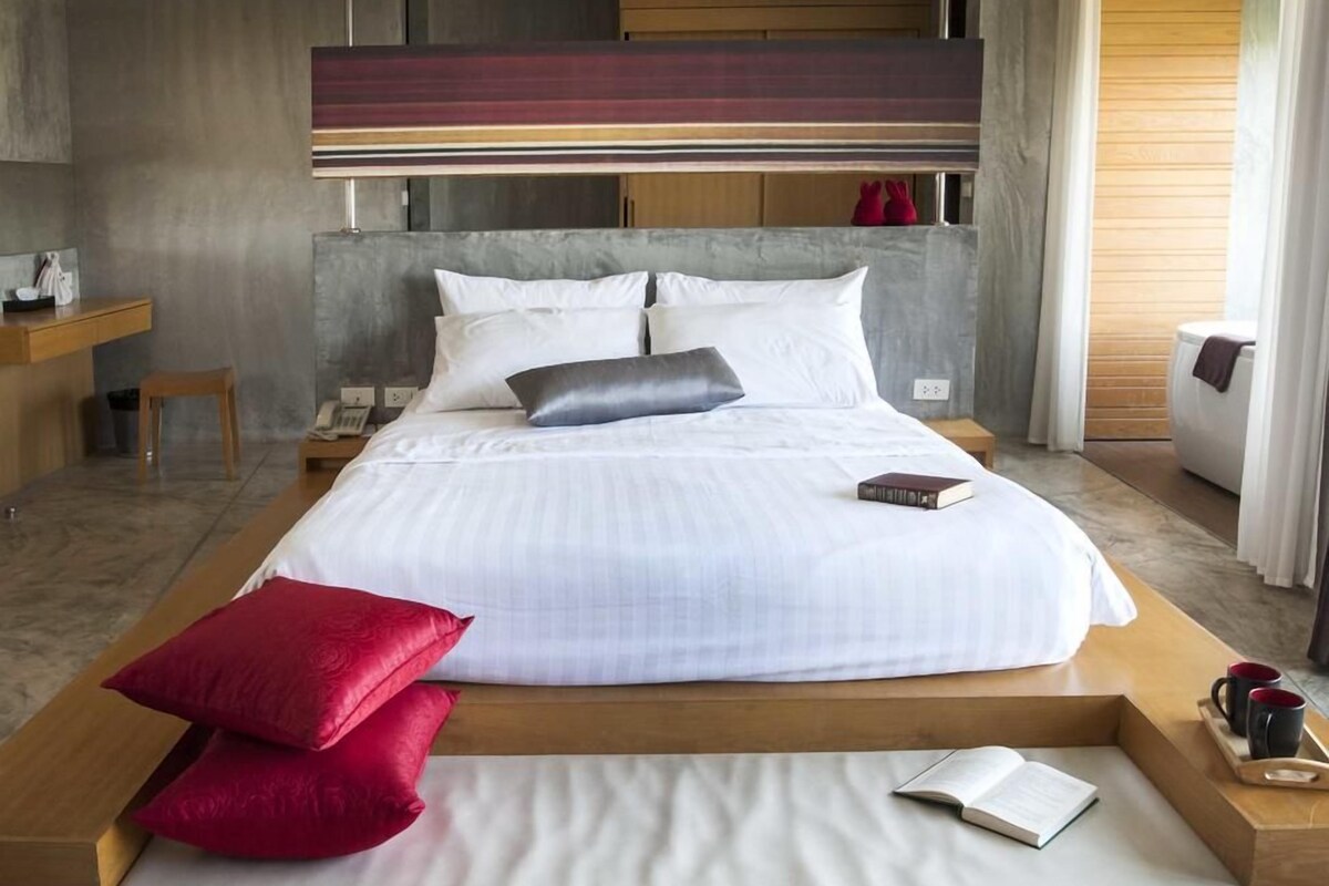 A modern hotel room with a large bed, a wooden bedside table, a small stool, a bedside phone, and a view of the outside.