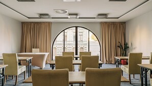 A hotel room with a large arched window, beige curtains, and a view of a building. There are multiple tables and chairs arranged for dining.