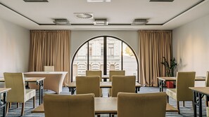 Quarto de hotel com uma grande janela arqueada, cortinas bege e vista para um edifício. Há várias mesas e cadeiras dispostas para refeições.