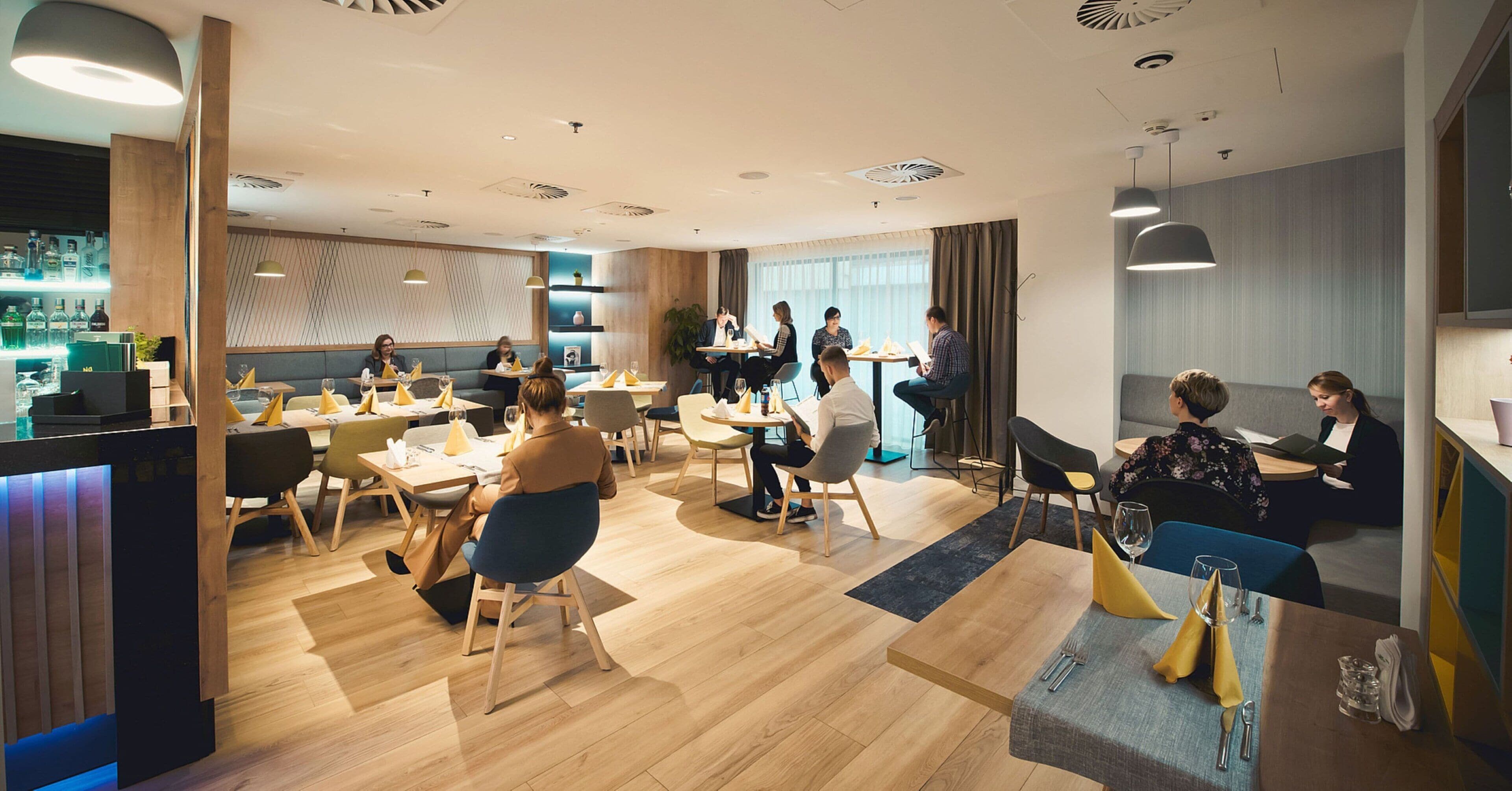 A modern dining area with wooden flooring, tables with yellow napkins, and a bar area with bottles.
