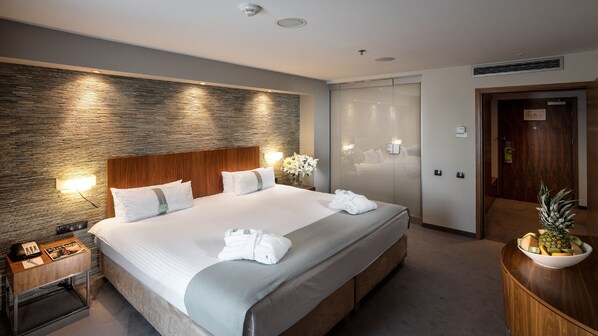 Quarto de hotel com uma cama grande, cabeceira de madeira, mesa de cabeceira com abajur, cômoda de madeira e uma cesta de frutas.
