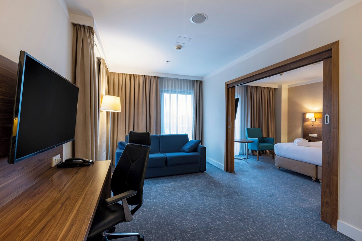 Quarto de hotel com televisão de tela plana, escrivaninha, sofá azul, cama e banheiro visível por uma porta aberta.