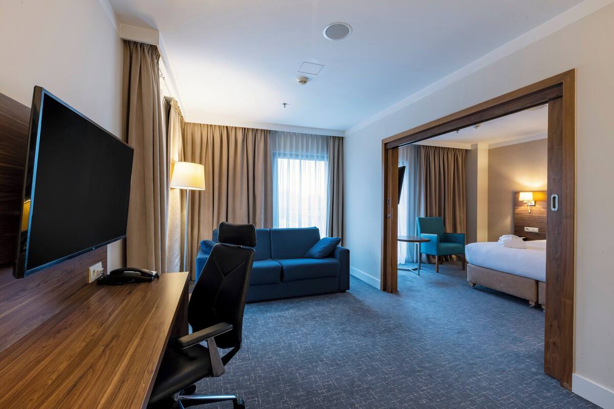 Habitación de hotel con televisión de pantalla plana, escritorio, sofá azul, cama y baño visible a través de una puerta abierta.