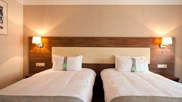 Habitación de hotel con dos camas individuales, cabecera de madera y mesitas de noche fijas a la pared con lámparas.
