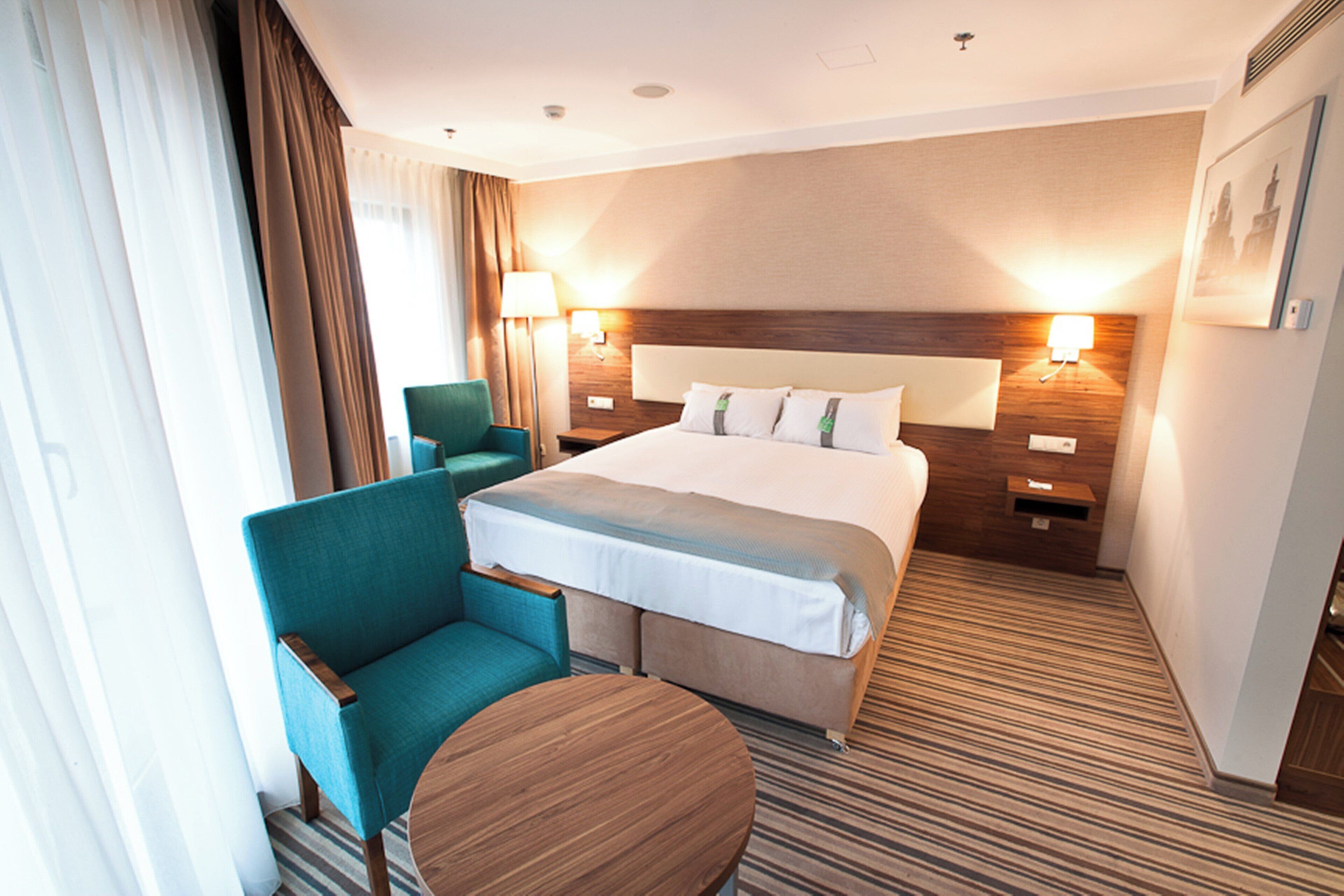 Habitación de hotel con una cama grande, un área de descanso con una silla y una mesa, y dos lámparas de noche.