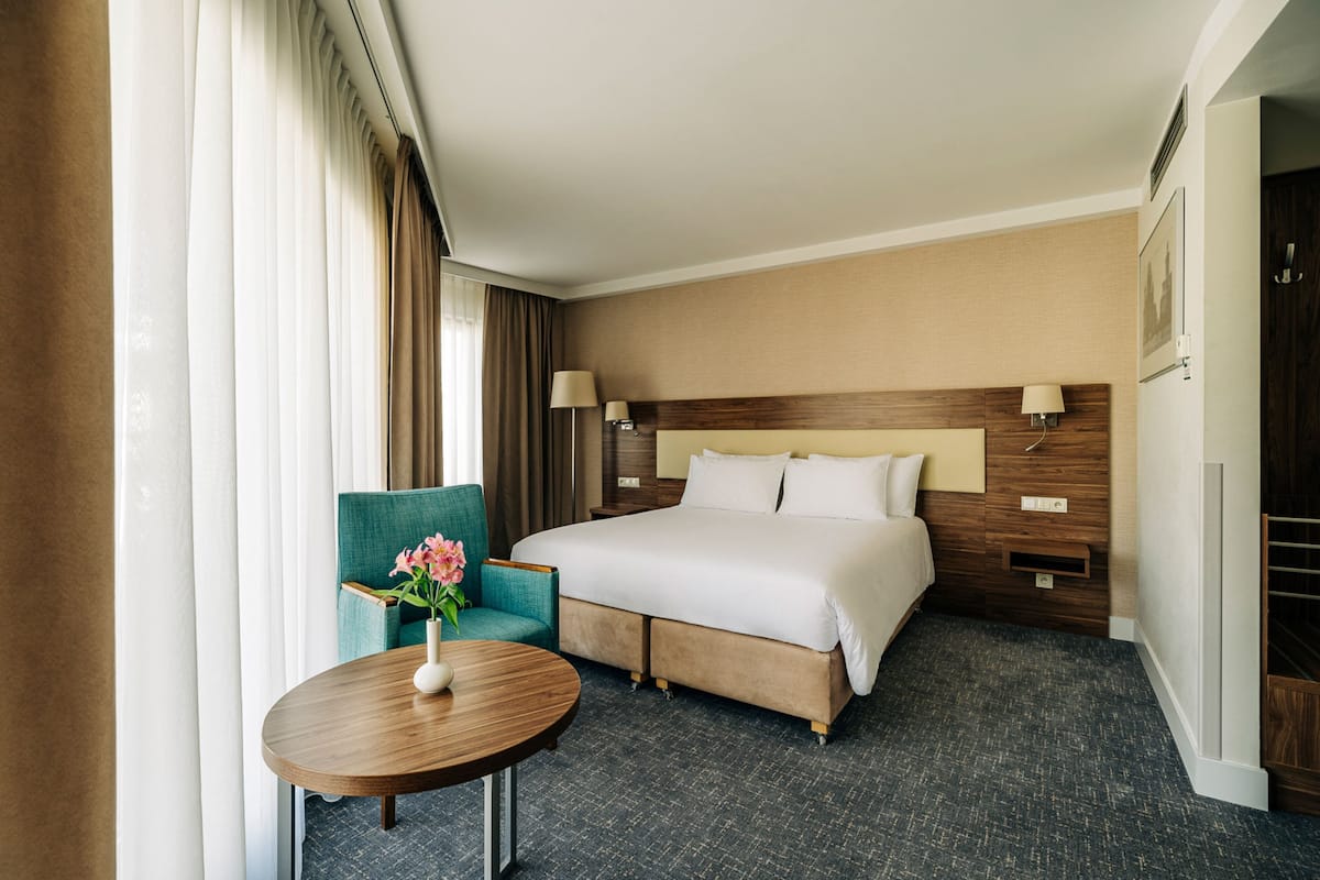 Una habitación de hotel con una cama grande, una mesa redonda con un jarrón de flores, un sillón color verde azulado y un cabecero de madera.