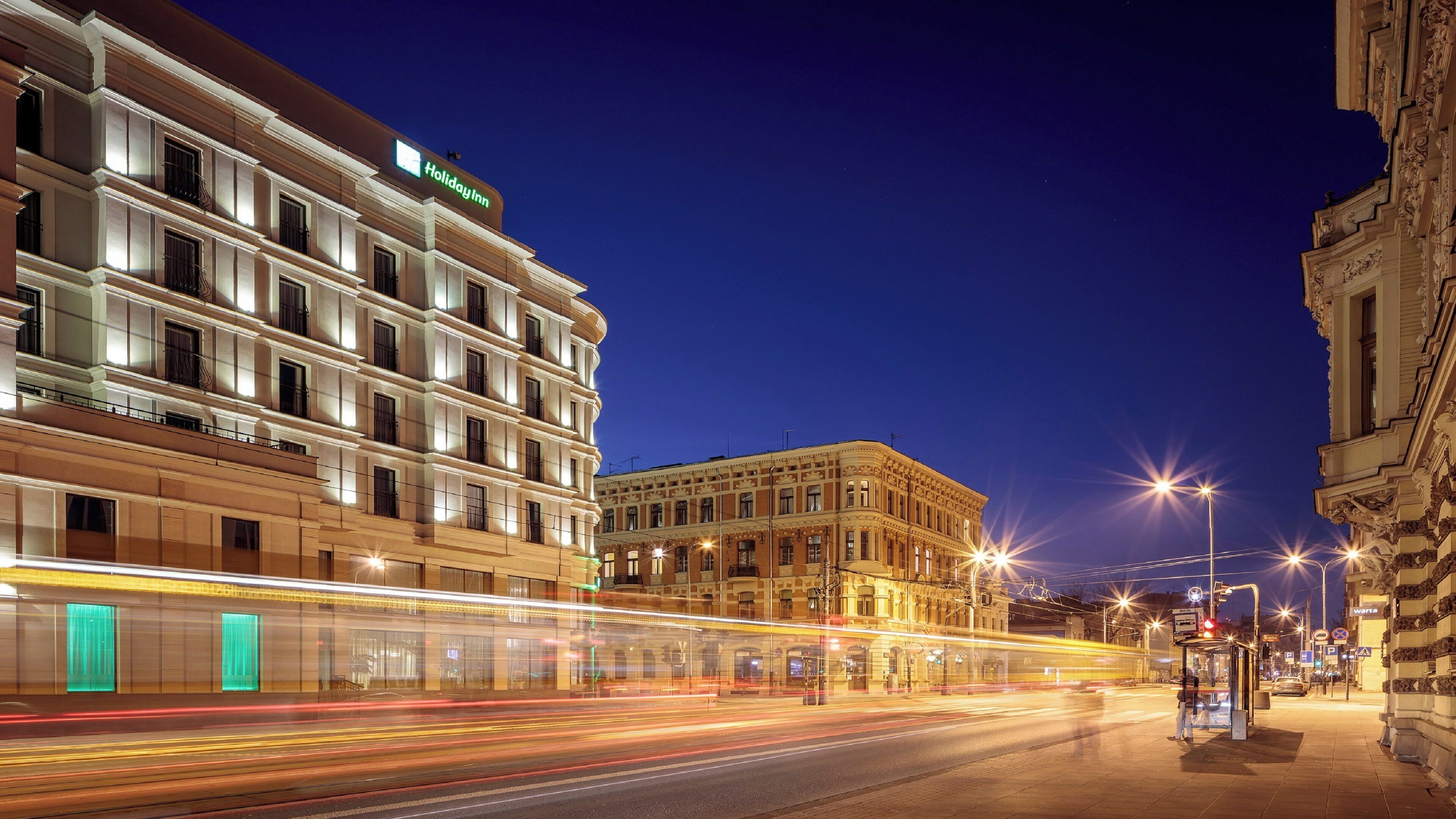 A multi-story building with a Holiday Inn sign, a street with light trails from moving vehicles, and a bus stop with a shelter.