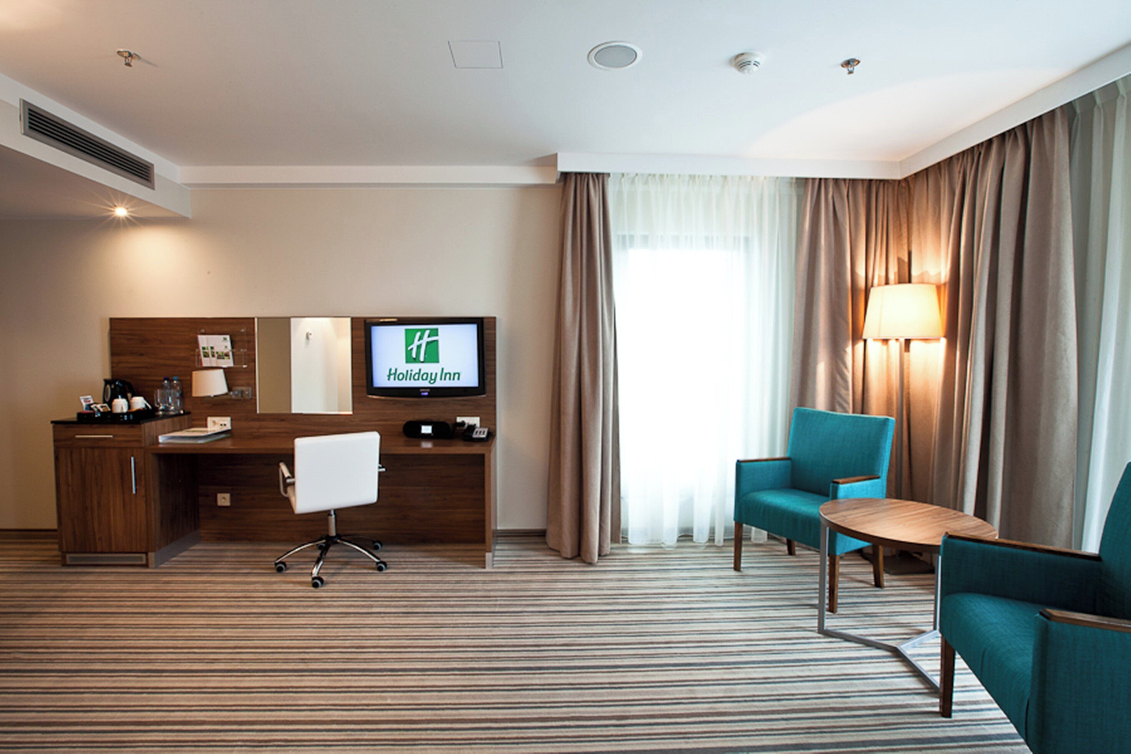 Quarto de hotel com escrivaninha, cadeira, televisão exibindo o logotipo Holiday Inn, luminária e cortinas.