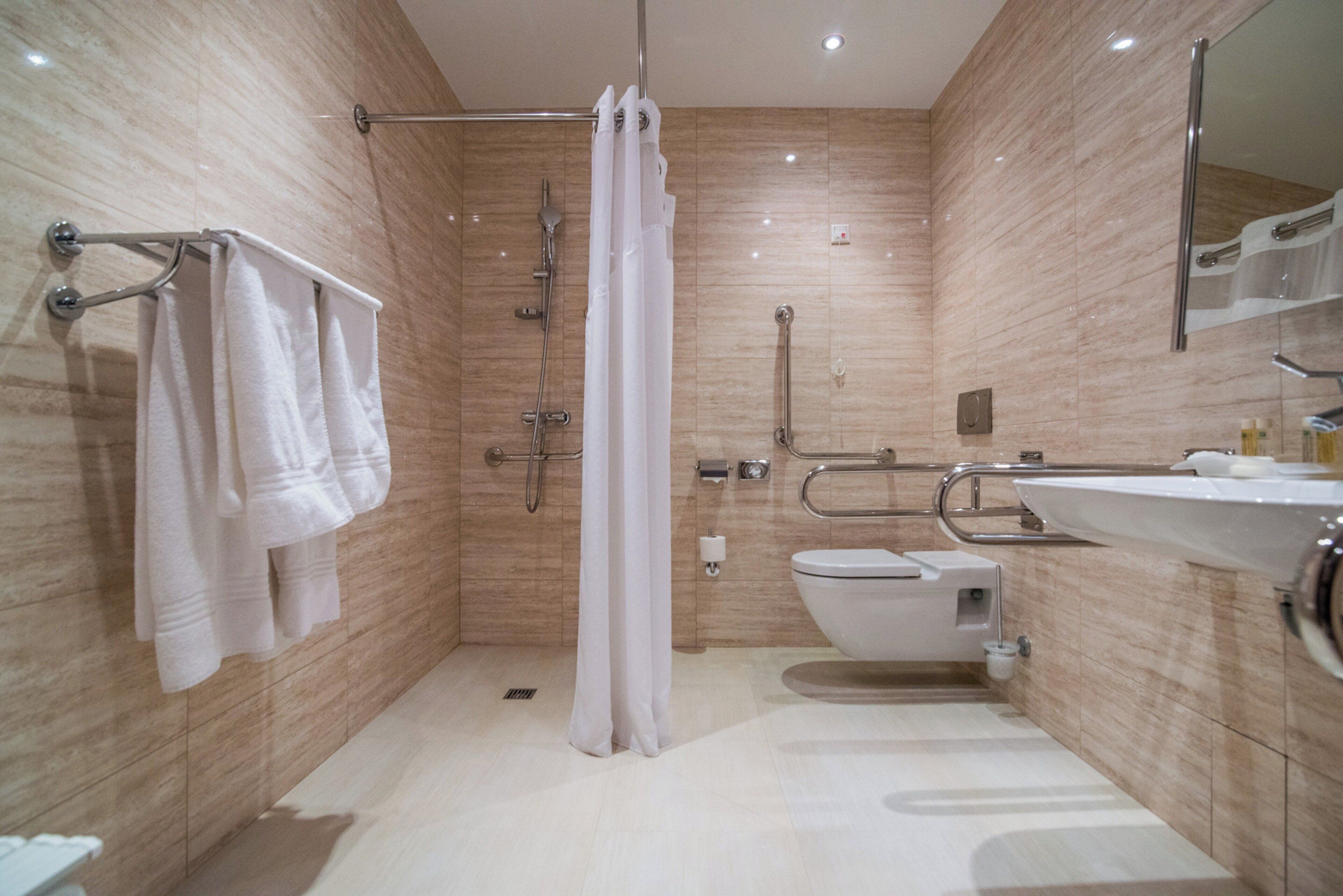 A modern bathroom with a shower, toilet, and sink, equipped with grab bars and a hand dryer.
